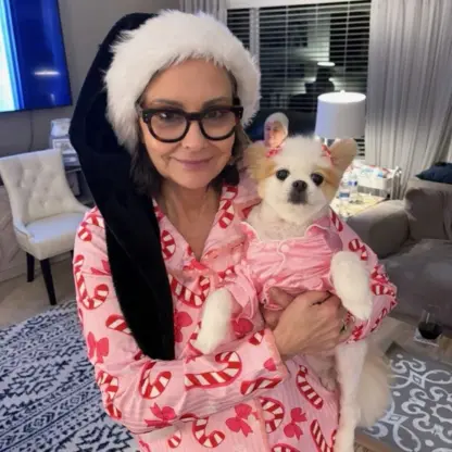 A woman in glasses and a black Santa hat, wearing pink pajamas with candy canes, holds a small dog dressed in matching pajamas in a cozy living room.