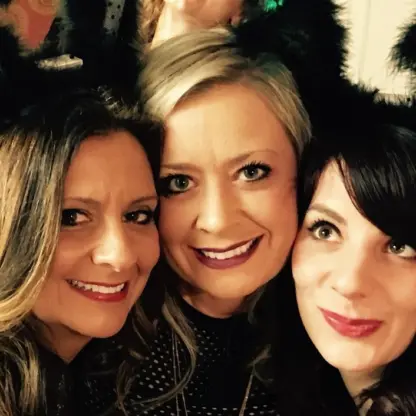 Three women smiling closely together, wearing black furry headbands with upright animal-like ears, posing for a selfie at what appears to be a festive indoor event.