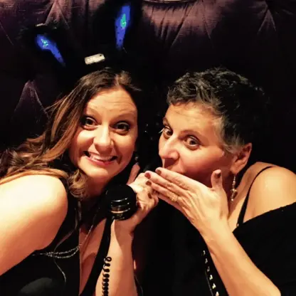 Two women smiling; one wears light-up bunny ears and holds a vintage phone, while the other covers her mouth as if sharing a secret. Both are in black outfits, seated against a tufted, dark purple background.