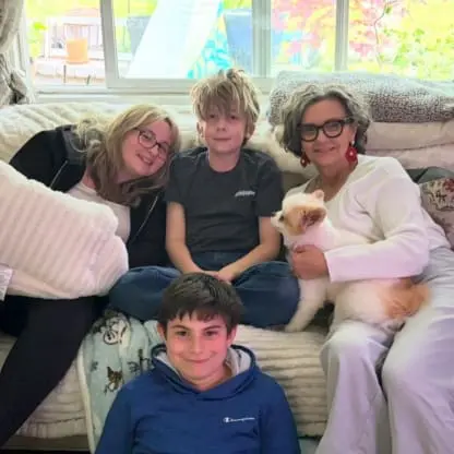 Four people sit on a couch, two adults and two kids, with a white dog on one adults lap. They are smiling, and sunlight shines through a window behind them, revealing greenery outside.