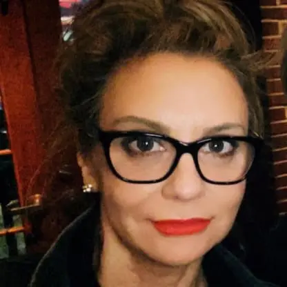 Woman with light brown hair in an updo, wearing black glasses, red lipstick, and a black top. She is indoors with a blurred background of a wooden door and brick wall.