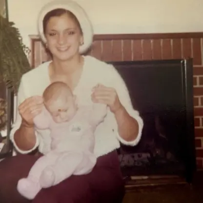 A woman wearing a white hat holds a baby dressed in pink while sitting in front of a brick fireplace. There is a potted plant and a vase of yellow flowers on a table beside them.