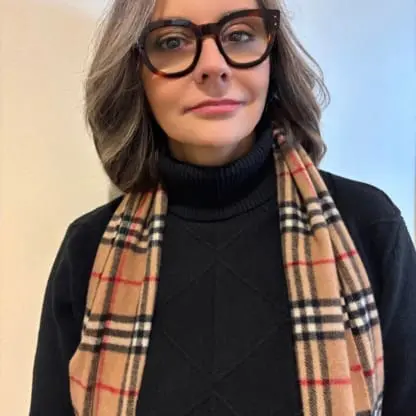 A woman with gray hair, wearing large glasses, a black turtleneck sweater, and a beige plaid scarf stands against a light background, looking slightly to the side with a neutral expression.