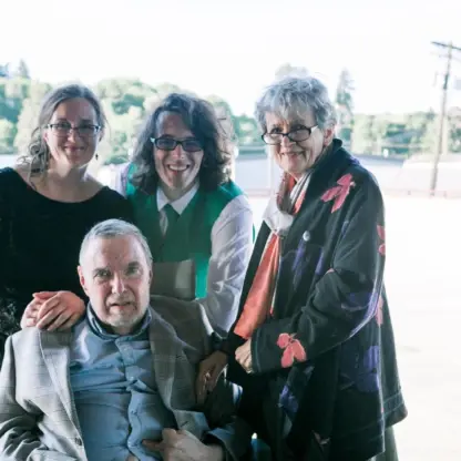 Four people pose together outdoors, three standing and one seated in a wheelchair. They are dressed semi-formally and smiling, with greenery and buildings visible in the bright background.