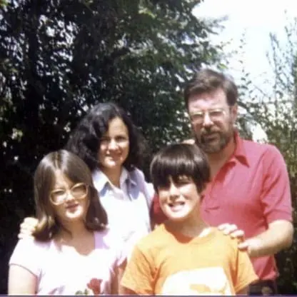 A man, woman, and two children stand outdoors, smiling. The man wears glasses and a red shirt. The woman has dark hair and wears a light shirt. The children wear glasses and casual clothes. Trees fill the background.