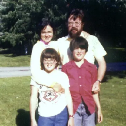 Four people stand outside on grass: two adults in back, one with glasses and a beard, and two children in front, both with glasses; trees and a road are in the background on a sunny day.