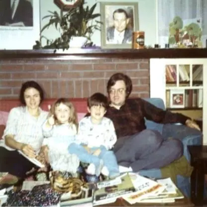 A family of four, two adults and two children, sit on a couch in a cozy living room with a brick fireplace, framed photos, houseplants, and magazines scattered on the table in front of them.