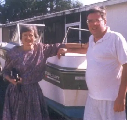 A woman in a purple dress and a man in a white shirt and shorts stand smiling next to a boat labeled STARCRAFT outside, with a building in the background.