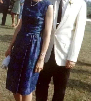 A woman in a blue dress and a man in a white jacket with sunglasses stand together on grass at an outdoor event, with people in the background.