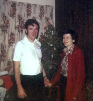A man in a white shirt and a woman in a red sweater stand holding hands in front of a small decorated Christmas tree, with floral curtains in the background.
