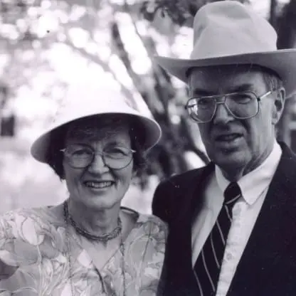 An older woman and man, both wearing glasses and hats, smile while standing outdoors. The man is in a suit and tie with a cowboy hat; the woman wears a patterned dress and light hat. The image is in black and white.