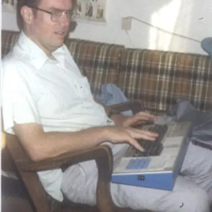 A man sits on a chair typing on a vintage computer keyboard placed on his lap, with a plaid couch and wall maps visible in the background.