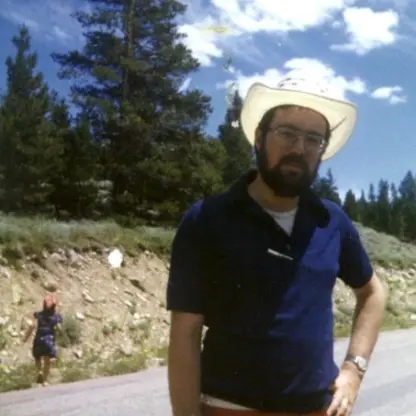 A bearded man wearing glasses and a white cowboy hat stands on a roadside with trees and blue sky in the background. A person in a red hat is walking in the distance.