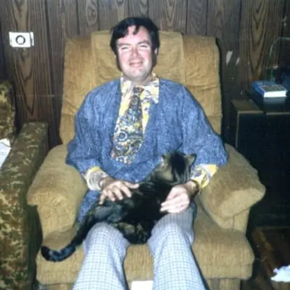 A smiling man in a patterned shirt and tie sits on a brown armchair, petting a tabby cat on his lap. Wood-paneled walls and books are visible in the background.