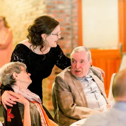 A woman in a black dress stands and smiles with her arms around an older woman and an older man seated at a table, both dressed formally, in a warmly lit room with exposed brick walls.