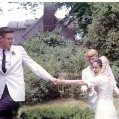 A bride and groom in white hold hands and smile outdoors, with two men in suits standing nearby. Green trees and a house are in the background.
