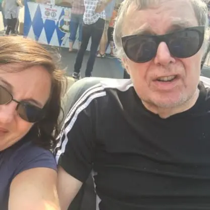 A woman and an older man, both wearing sunglasses, smile at the camera outdoors. The man wears a black Adidas shirt; people and a food stand are visible in the sunny background.