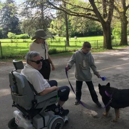 An older man in a wheelchair, a woman, and a boy holding a German Shepherd on a leash are outdoors on a road next to a park with trees and a fence.