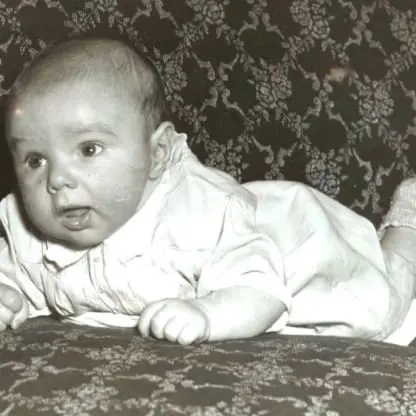 A baby wearing a light-colored outfit and socks lies on their stomach on a patterned couch, looking slightly to the left with a curious expression. The photo is in black and white.