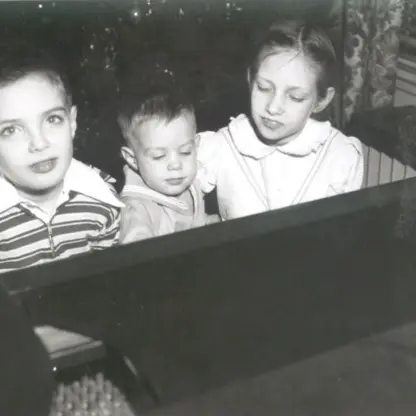 Three children sit closely together at a piano, with one older child looking at the camera, the youngest looking down, and another child watching the youngest. The scene is indoors, in black and white.