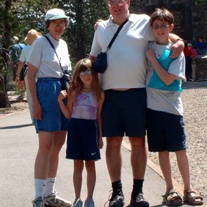 A family of four stands outdoors on a sunny day, smiling at the camera. Trees and a stone building are in the background. The family members wear casual summer clothes and sunglasses.