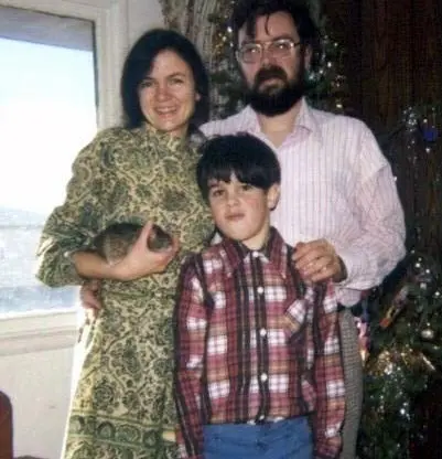 A woman in a green dress, a man in glasses, and a boy in a plaid shirt stand together indoors by a window and a decorated Christmas tree. The woman is holding a small animal. All are smiling.