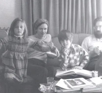 A black-and-white photo of a family sitting on a couch. Two children and two adults pose together, with books and papers spread out on the table in front of them. Curtains hang in the background.