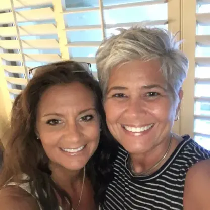 Two women smiling together in front of white window shutters. One has long brown hair and is wearing a white top; the other has short gray hair and a black-and-white striped sleeveless top.