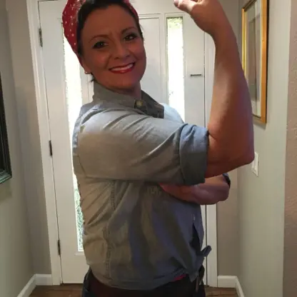 A person wearing a red bandana and denim shirt poses with a flexed arm, smiling confidently in a hallway, reminiscent of the We Can Do It! Rosie the Riveter poster.
