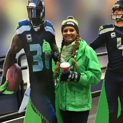 A woman in Seahawks gear stands between cardboard cutouts of two Seattle Seahawks football players, smiling and holding a coffee cup.