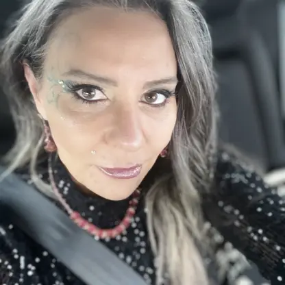 A woman with long gray hair, wearing a black sparkly top, pink earrings, and a necklace, sits in a car seat with a seatbelt on. She has facial makeup and a small piercing above her lip.