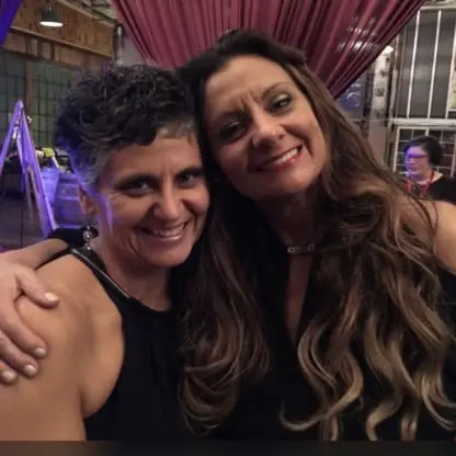 Two women smile and embrace at an indoor event with draped curtains and warm lighting. Other people and rustic decor are visible in the background.