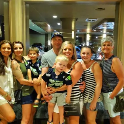 Seven people—four women, two young boys, and a man—pose and smile indoors near a large window. Most are dressed in summer clothes with stripes or shorts. Its a casual, cheerful group photo.
