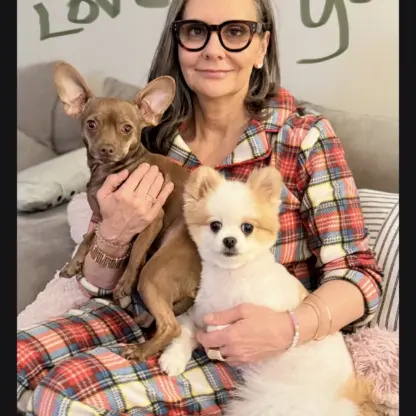A woman in plaid pajamas sits on a couch holding a small brown dog and a fluffy white dog. Love you is written on the wall behind her.
