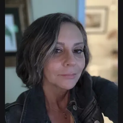 A woman with short brown and gray hair wearing a denim jacket, posing for a selfie indoors with artwork and soft lighting in the background.