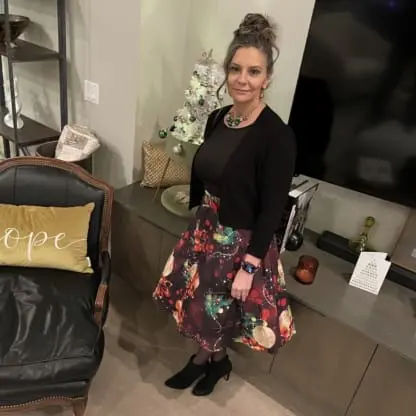 A woman in a festive skirt, black top, and boots stands beside a small decorated Christmas tree in a modern living room with a hope pillow on a chair.