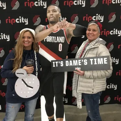 Two women pose with a life-size cutout of a Portland Trail Blazers basketball player. One holds a clock prop, the other holds a sign that says ITS DAMES TIME. The backdrop features repeated ripcity logos.