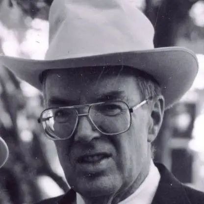 An older man wearing large glasses and a white cowboy hat, dressed in a suit, looks slightly past the camera in this black and white photo. Trees and blurred backgrounds are visible behind him.