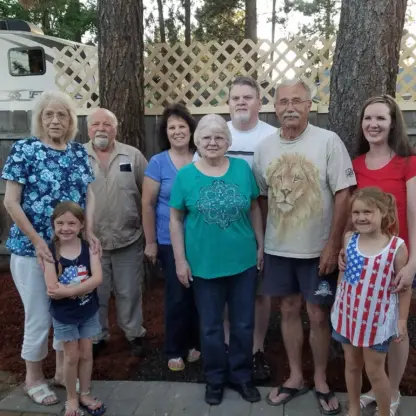 A group of eight people, including two young girls, posing for a photo outdoors in front of a lattice fence and pine trees.
