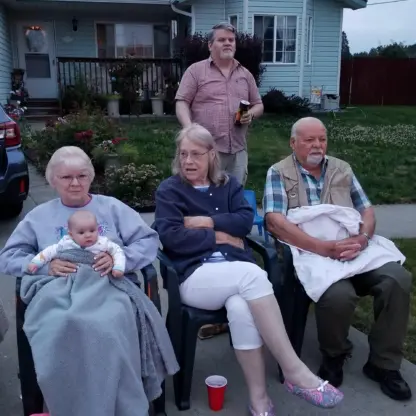 A group of four adults and a baby sitting on chairs in a driveway in front of a house.