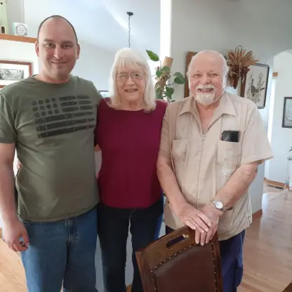 A younger man, an older woman, and an older man with a white beard stand together smiling inside a home.