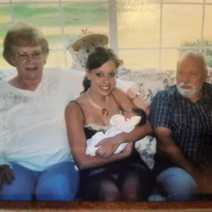 An older woman, a younger woman holding a newborn baby, and an older man sitting together on a couch in front of a window.