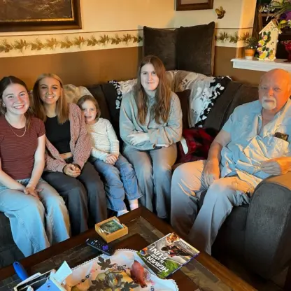 Four young women and an older man sitting together on a sectional sofa in a living room.
