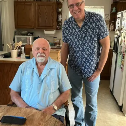 An older man with a white beard sits at a wooden table while a taller man stands beside him in a kitchen.