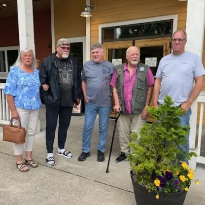 Five people standing together on a porch in front of a building entrance with a potted plant in the foreground.