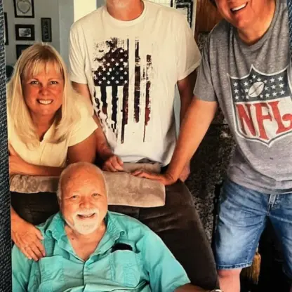 A family of four posing for a photo, with an older man sitting in a chair in front and a woman and two men standing behind him.