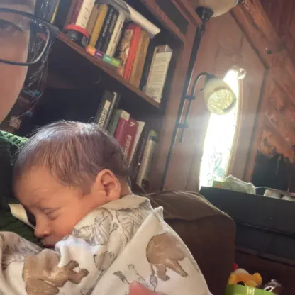 A person wearing glasses holds a sleeping newborn baby against their chest.