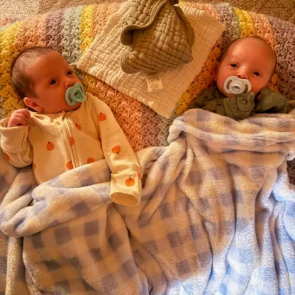 Two newborn twins lying side by side on a colorful crocheted blanket, both with pacifiers and covered by a blue and white checkered blanket.