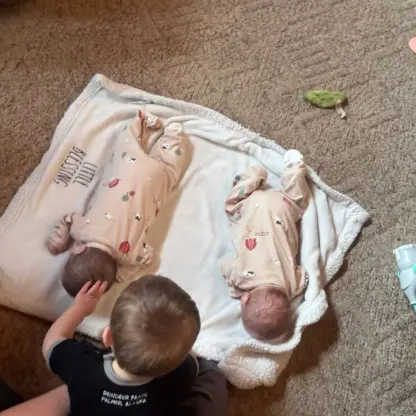 Two infants in patterned onesies lying on a white blanket as a toddler reaches out to touch one of them.