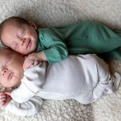 Two sleeping newborns lying on a textured white blanket, with the baby in green resting on the baby in white.